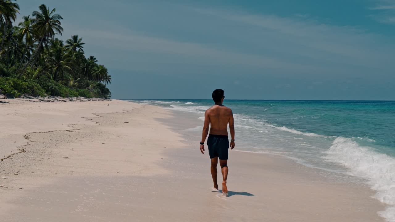 A solitary man walks along the untouched shoreline of Geiymiskih Fannu Beach in Fuvahmulah, surrounded by swaying palm trees and the gentle rhythm of turquoise waves.