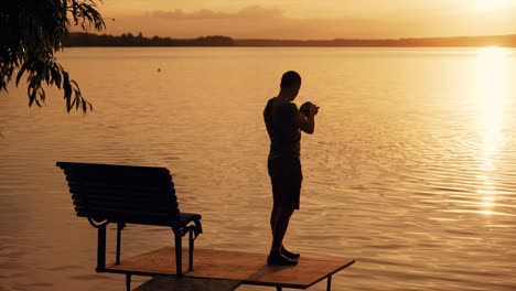 a talented photographer with a camera in his hands taking pictures of the sunset on the river on the bridge with a bench. silhouette of man with a camera on his favorite work.