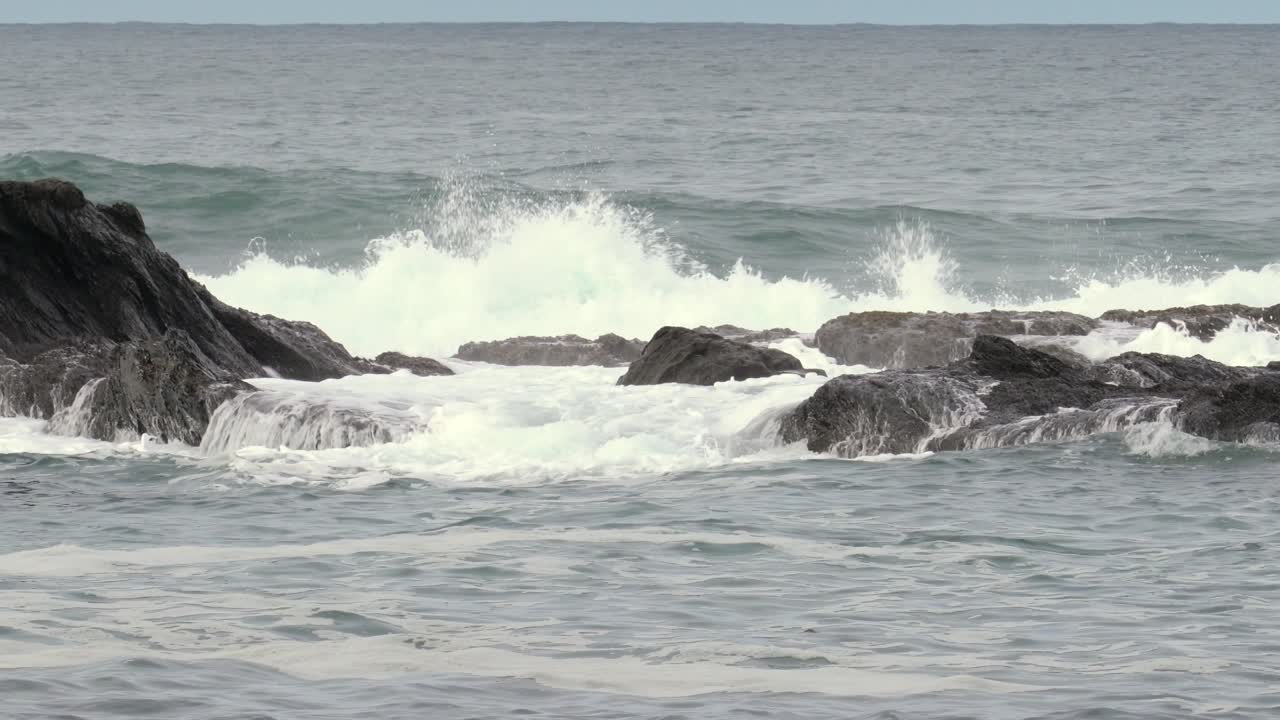 Waves crash against dark rocks in a serene ocean setting, creating splashes and white foam under a cloudy sky, capturing the power and beauty of nature