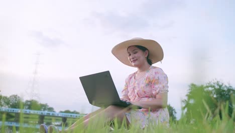 Beautiful Asian woman in hat sitting on lawn working on computer feeling successful