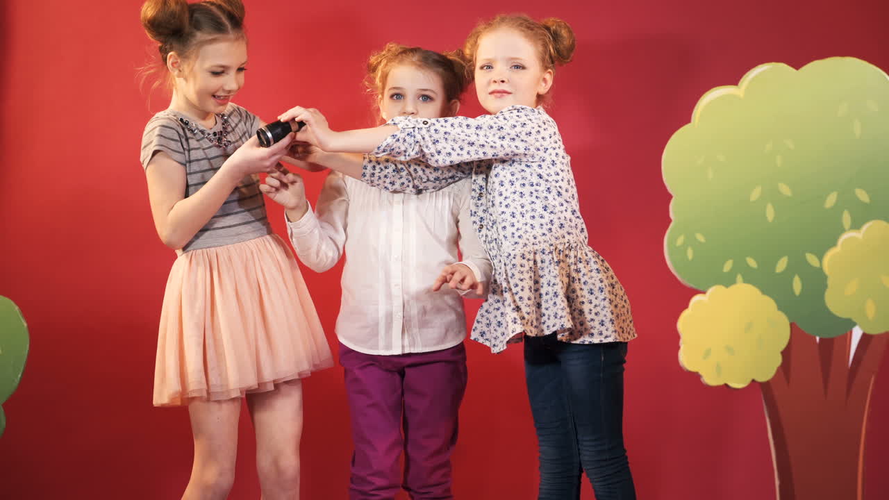 Children are playing with binoculars and looking for something on the red studio background. Three cute girls posing to camera near the tree decoration.
