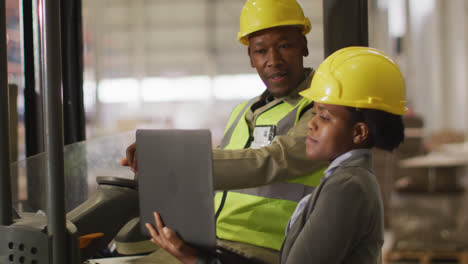 african american male and female workers wearing safety suits and using laptop in warehouse