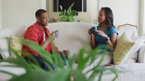 Feliz-Pareja-Birracial-Sentada-En-El-Sofá-Con-Café-Y-Hablando