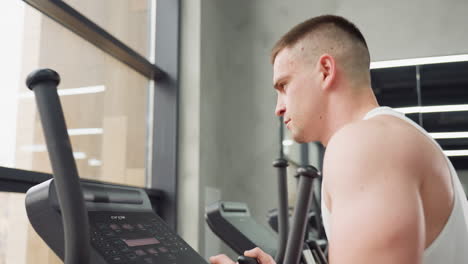focused sports man exercising on elliptical trainer in bright modern gym with large window and mirror reflecting workout area and equipment while concentrating intensely on cardio session momentum