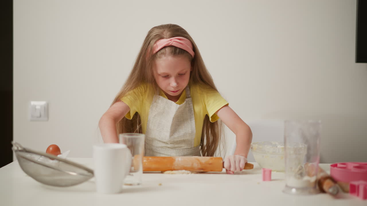 Young girl wearing yellow shirt and apron rolling dough with strength and speed on white kitchen table during baking session, focused on preparing dough with surrounding baking tools