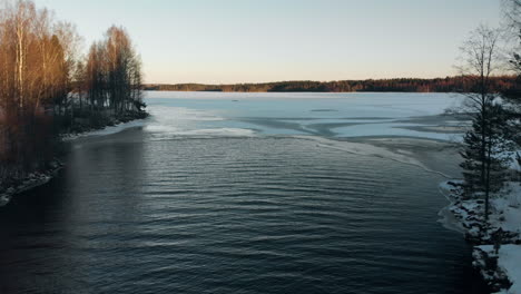 aerial, reversing, drone shot, above a river, of first ice on a lake, surrounded by leafless forest and earley snow on the ground, on a sunny, autumn day, near joensuu, north karelia, finland