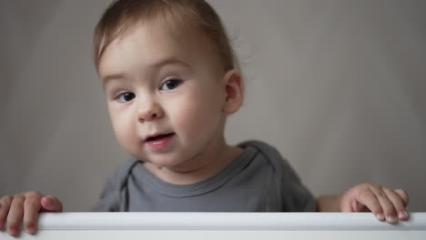 cute adorable kid wearing grey shirt stands in cot holding by the handrail. nice child sits in his crib hiding behind the bed border.