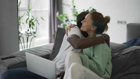 feliz pareja biracial sentada en el sofá con una computadora portátil, felicitándose entre sí