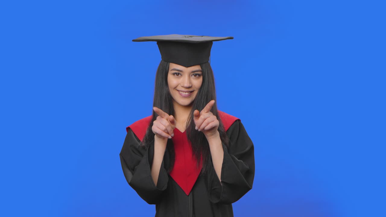 Ritratto di studentessa in abito da laurea sorridente, che forma un cuore con le dita e poi manda un bacio. Giovane bruna in posa su sfondo blu. Primo piano. Pronto per slow motion a 59.94fps