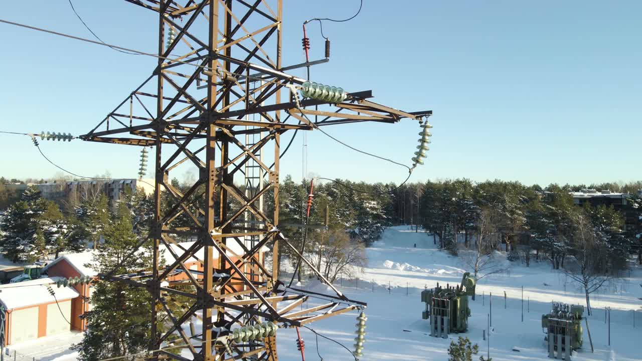 Drone rise reveals lattice tower, suspension insulators, and nearby substation across sunlit snowy landscape