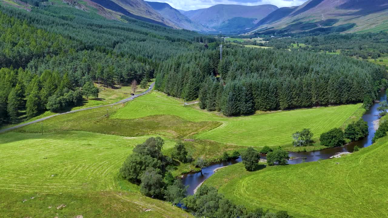 Drone glides above green river valley, dense forest, and distant mountains under bright daylight