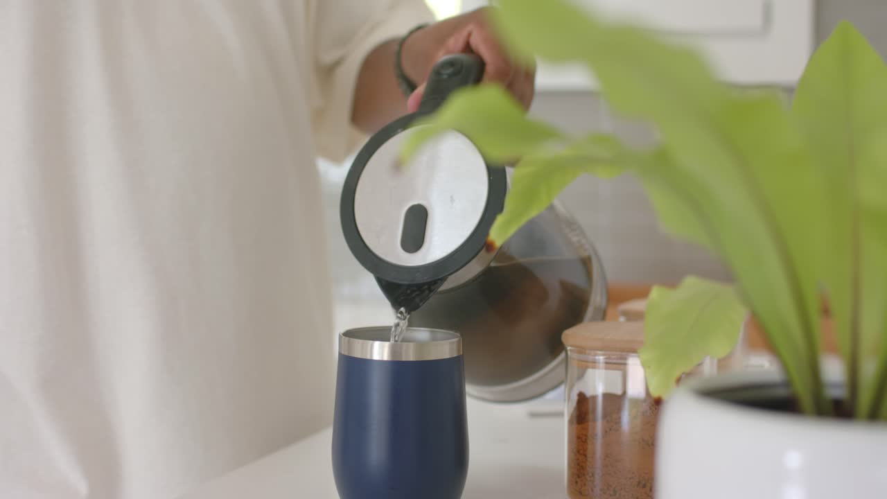 Uomo africano che solleva una caraffa, versando in una tazza da viaggio blu, riempiendola sul bancone della cucina.