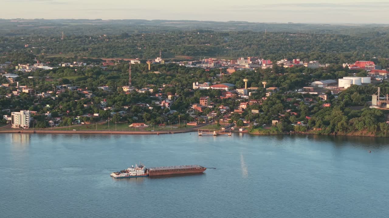 Grain barge navigates the Paraná River before the urban skyline of Encarnación, Paraguay, captured in a smooth drone orbit showing the riverside, buildings, and distant forested hills