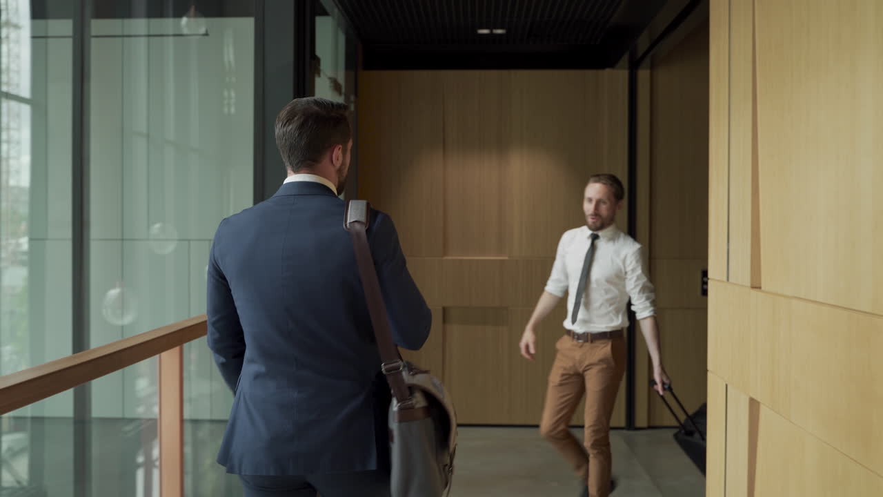 An attractive young businessman walks through a corridor with a man bag. He meets a colleague, greets him and they start talking.