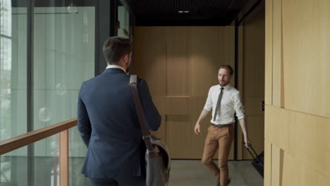 An-attractive-young-businessman-walks-through-a-corridor-with-a-man-bag.-He-meets-a-colleague,-greets-him-and-they-start-talking.