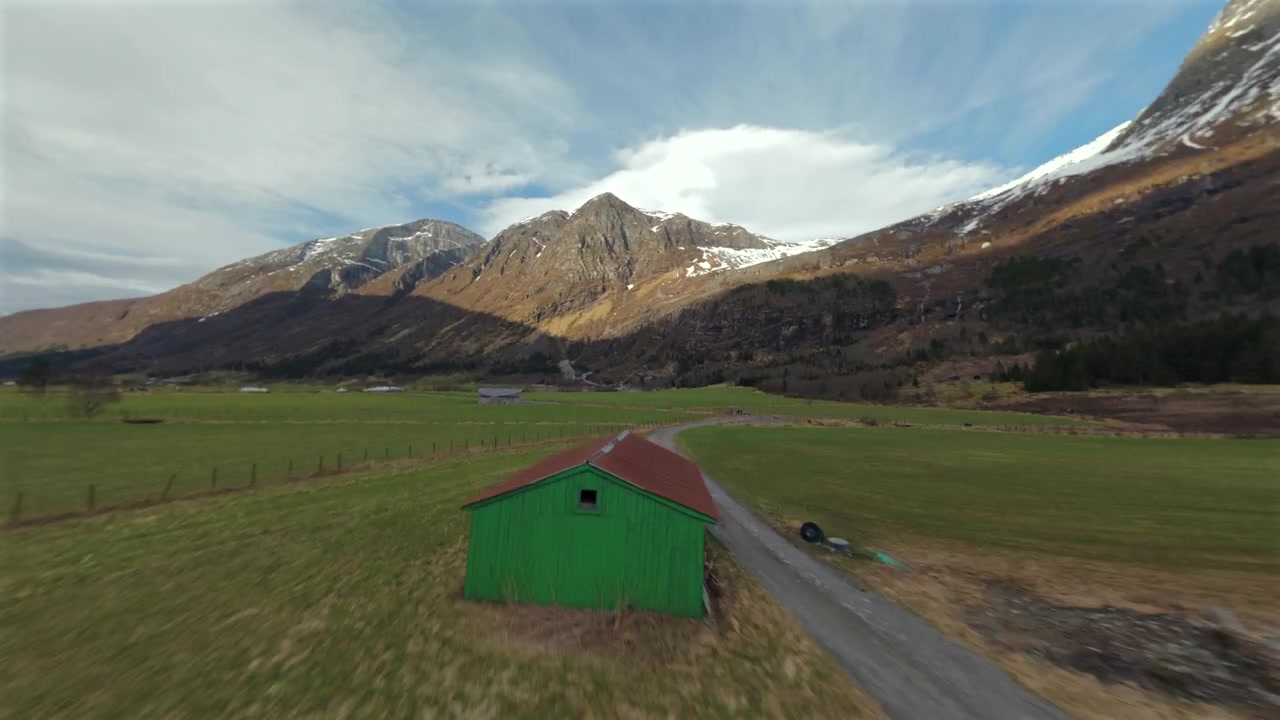 Paesaggio di valle montana in Norvegia catturato da un drone FPV con cime rocciose, campi verdi e chiazze di neve sotto la luce dell'ora d'oro.