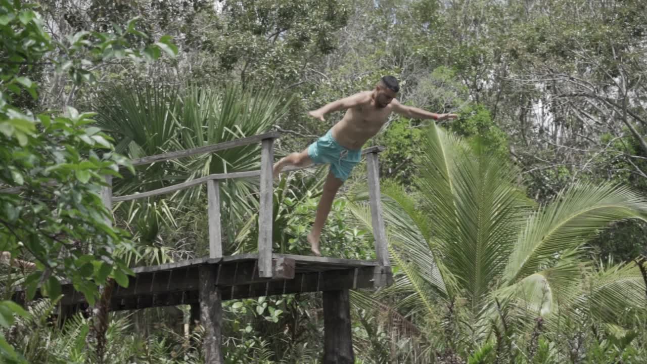 A dynamic shot capturing a man in light blue swim trunks as he jumps from a wooden diving platform into a cenote (natural sinkhole) in the Tulum region of Mexico