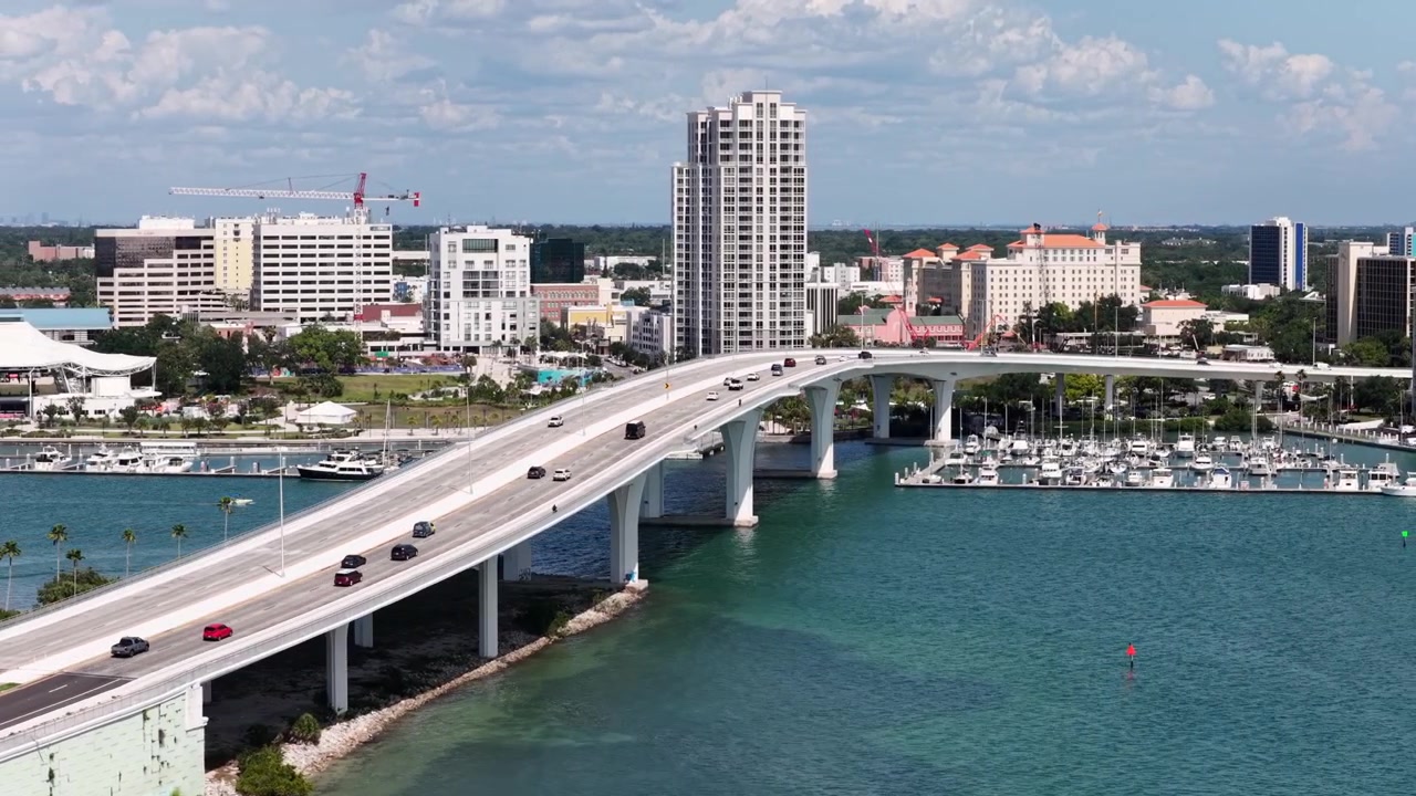 Auto percorrono un ponte sopraelevato che collega il centro di Clearwater, Florida, a un porto turistico pieno di barche, un grattacielo si erge sopra lo skyline vicino alle acque turchesi della baia. Ampia ripresa aerea con drone