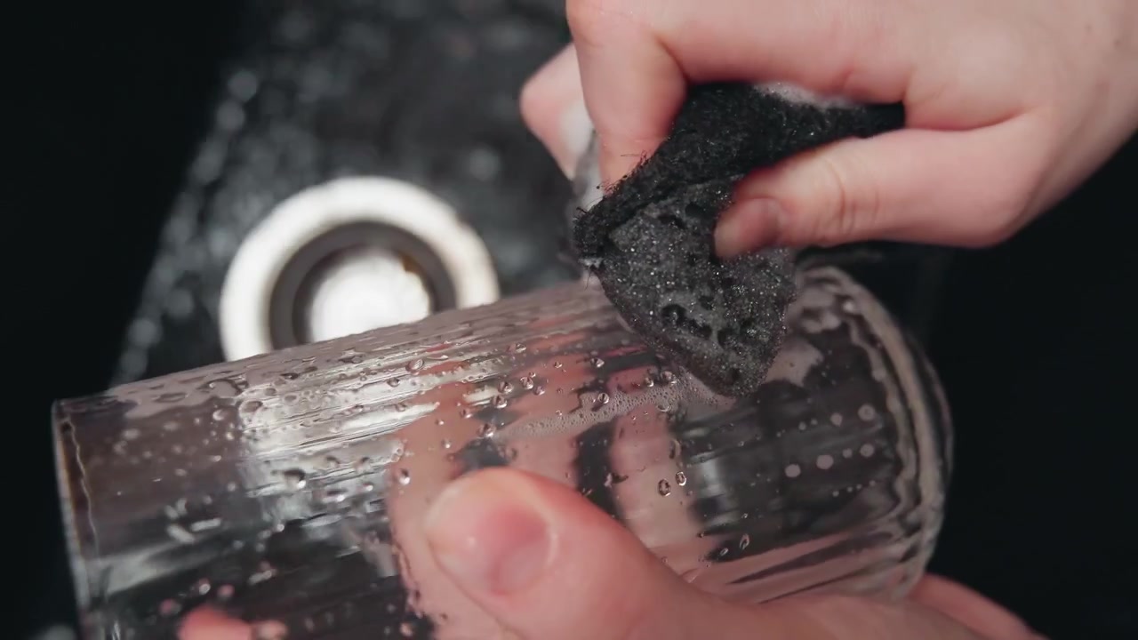Close macro side view shows hands cleaning interior of clear plastic cup with dark abrasive sponge, producing rotational and downward scrubbing strokes, wet streaks, and soap bubbles