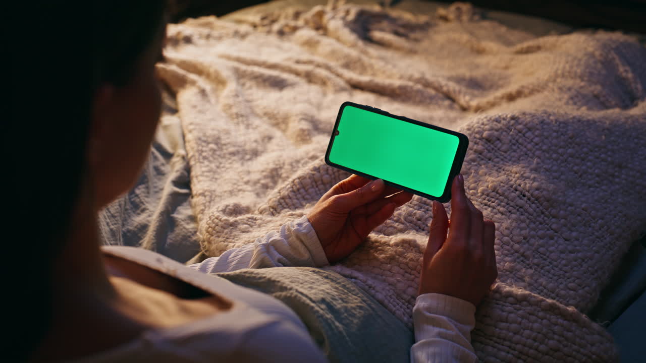 Primo piano di una donna che guarda un video su uno smartphone a letto. La donna guarda uno schermo verde.