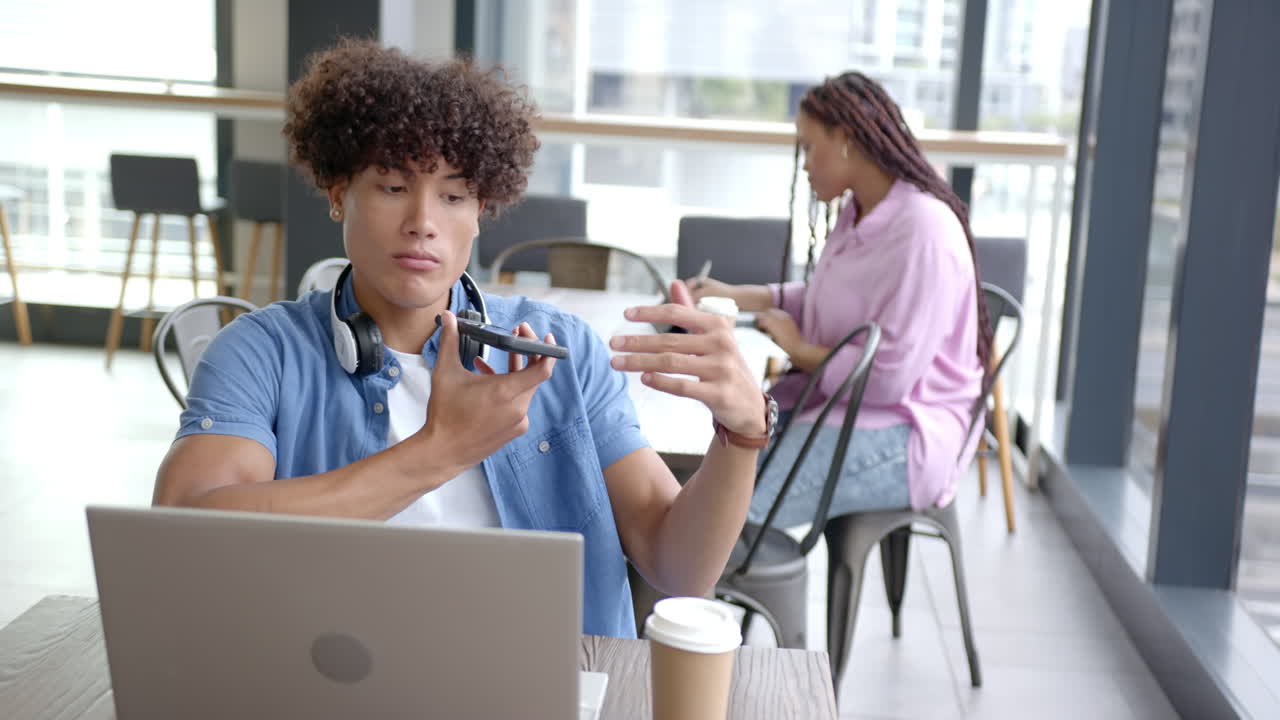 Using smartphone for voice message, man working on laptop in cafe with coffee