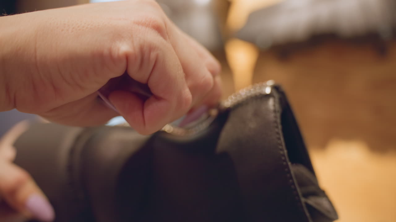 Closeup of hands with lilac manicure testing zipper on black leather chukka boot, sliding puller up and down verifying smooth glide, assessing build quality for purchase decision under warm lighting