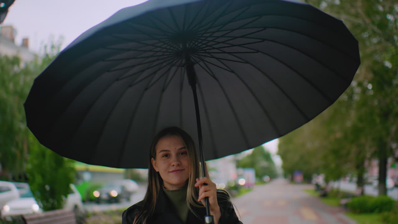 검은색 우산 뒤에 숨어 우산을 머리 위로 펼쳐 빙글빙글 돌리는 멋진 여성의 모습이에요. 흐린 날, 자동차, 나무, 건물이 흐릿하게 보이는 도시 거리에 서 있어요.