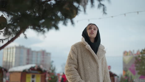 girl in warm beige winter jacket with black hood walks through snowy street, gazing up at a beautifully decorated tree, soft lights create a cozy atmosphere, with blurred festive background