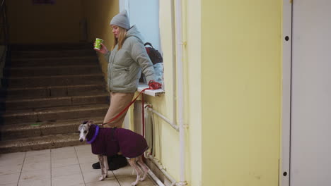 woman in winter clothing drinking from green cup while standing near indoor stairway holding red leash of whippet dressed in purple jacket with purple toy, both waiting in casual quiet urban moment
