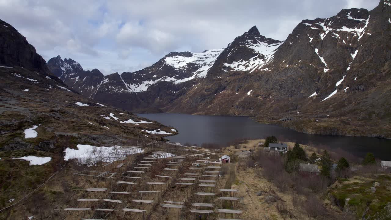 disparo ascendente de un avión no tripulado de una vista hacia un lago y un estante de pescado seco con montañas cubiertas de nieve a finales de abril, en lofoten