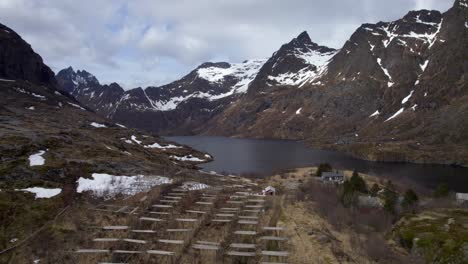 Toma-Ascendente-De-Un-Dron-De-Una-Vista-Hacia-Un-Lago-Y-Un-Estante-De-Pescado-Seco-Con-Montañas-Cubiertas-De-Nieve-A-Finales-De-Abril,-En-Lofoten