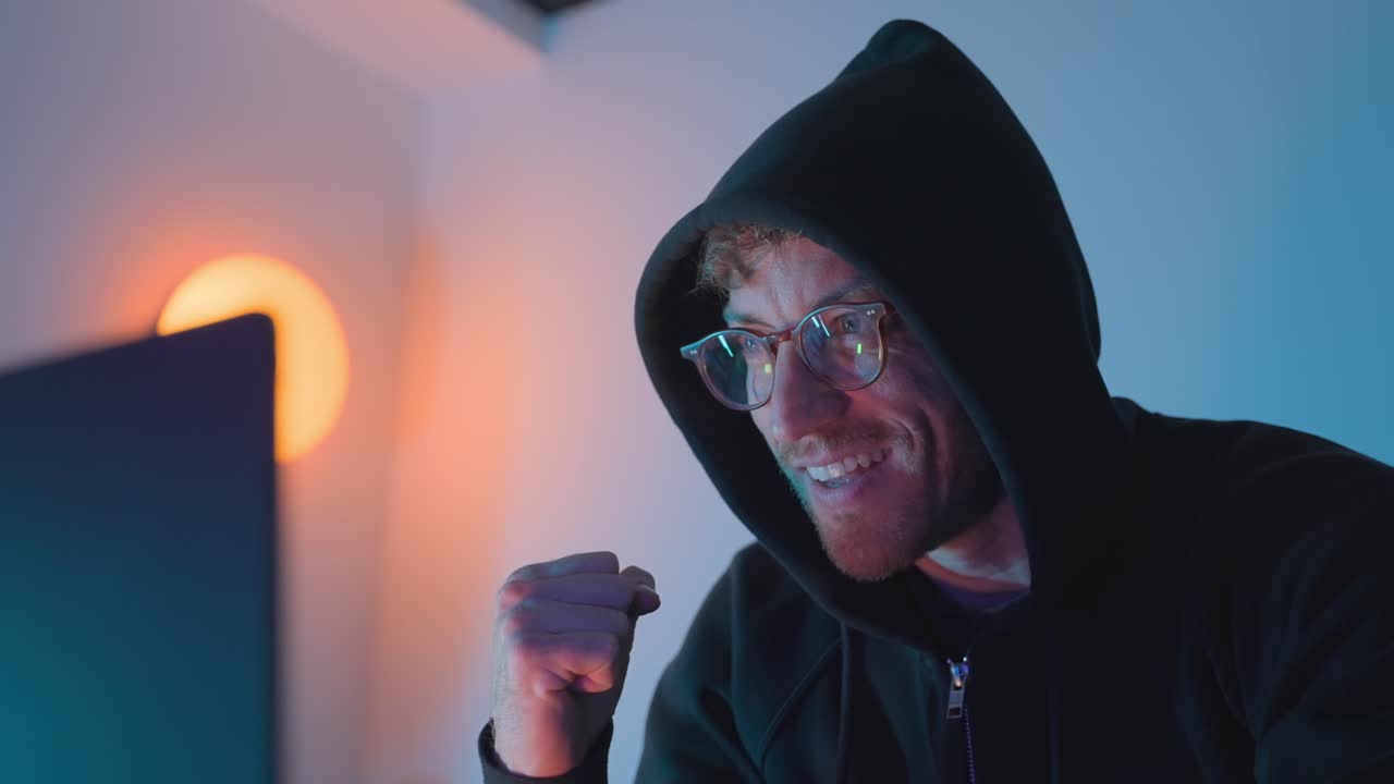 Hacker ved bærbar computer. Hætteklædt mand fejrer ved computeren i blåt lys, hæver knyttet hånd med et begejstret udtryk. Koncept for hacking, cyberkriminalitet, databrud, malwareangreb og cybersikkerhedstrusler.
