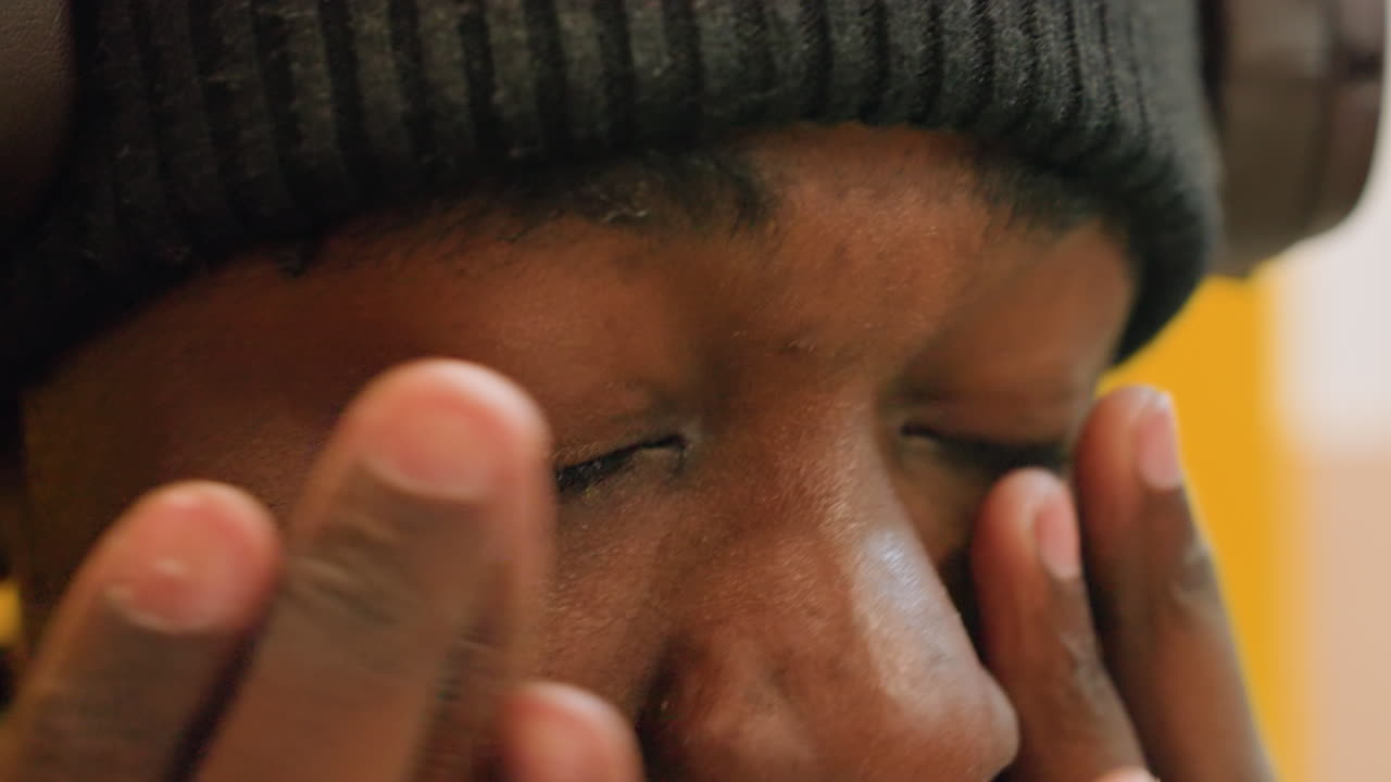 Close-up de homem negro esfregando os olhos, gorro e fones de ouvido, estudante noturno e produtor musical, fadiga e estresse, suor na testa, expressão reflexiva, fundo amarelo, luz de estúdio, brilho da tela, momento