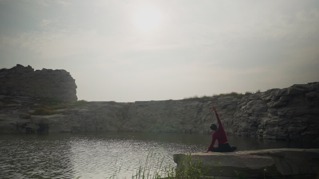 Woman practicing yoga by tranquil quarry lake in the morning sun with soft reflections on water, slow motion