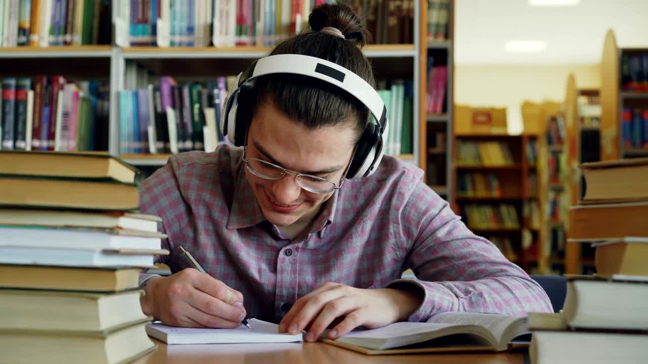 joven estudiante caucásico guapo sentado en la mesa en la biblioteca de la escuela grande está escribiendo algo en el cuaderno. está sonriendo y parece positivo y feliz, libros tendidos en la mesa
