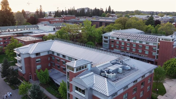 Edifici universitari in mattoni rossi con tetti in metallo, viali alberati, studenti sui sentieri e collina boscosa sotto la morbida luce della sera. Eugene, Oregon, USA. Ripresa aerea in sorvolo. Scena tranquilla.