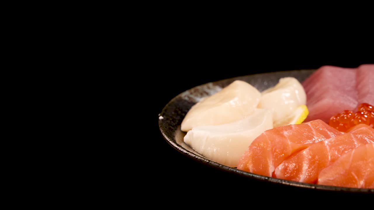 Đĩa sashimi tổng hợp gồm cá hồi, cá ngừ, sò điệp và trứng cá hồi xoay nhẹ nhàng trên nền đen dưới ánh sáng studio rực rỡ