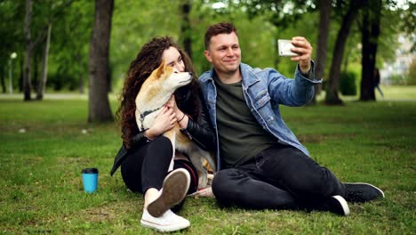 un joven guapo se está tomando una selfie con su novia que tiene un perro de pedigrí, el chico está besando a la chica y luego viendo fotos en la pantalla, la gente se está riendo.