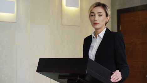 Close-up-view-of-caucasian-businesswoman-wearing-formal-clothes-on-a-podium-and-speaking-at-a-conference-in-front-of-many-people