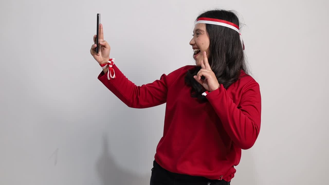 Smiling young Asian woman taking selfie using smartphone with make peace sign. Indonesian independence day celebration concept