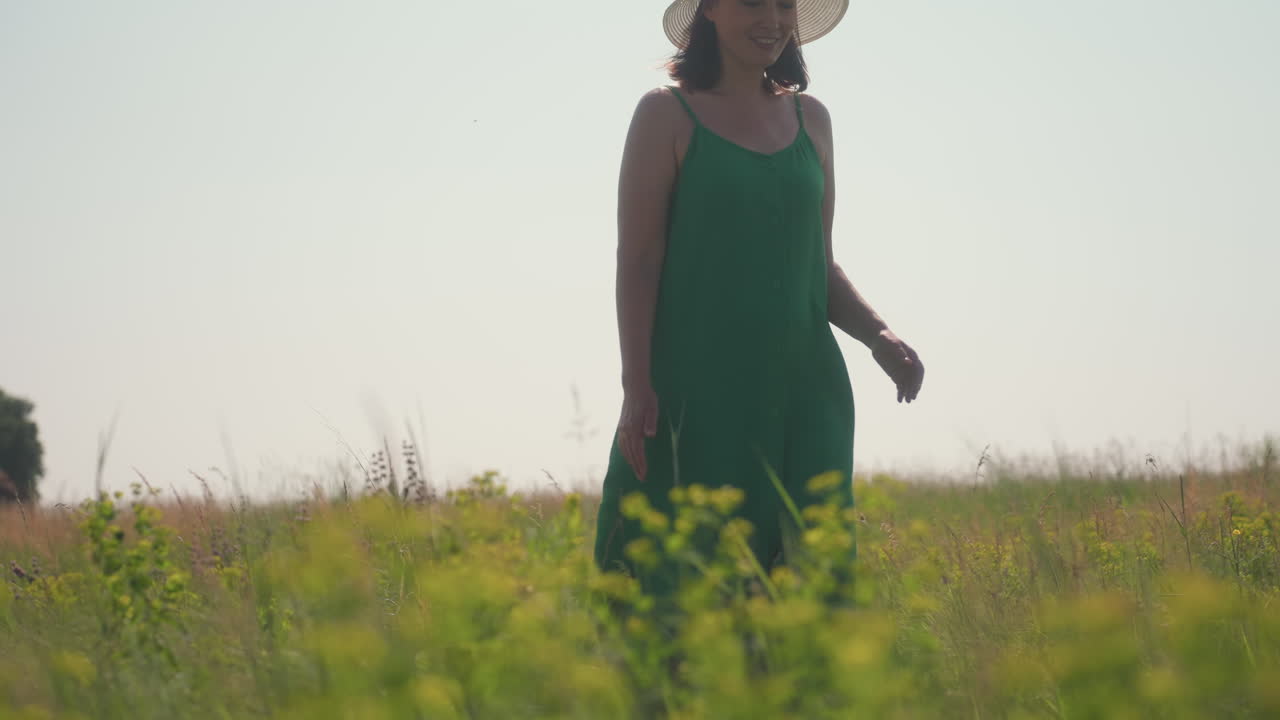 Woman in green dress and white hat walks gently through tall grassy field enjoying warm afternoon sun, surrounded by wildflowers and golden light under peaceful clear sky