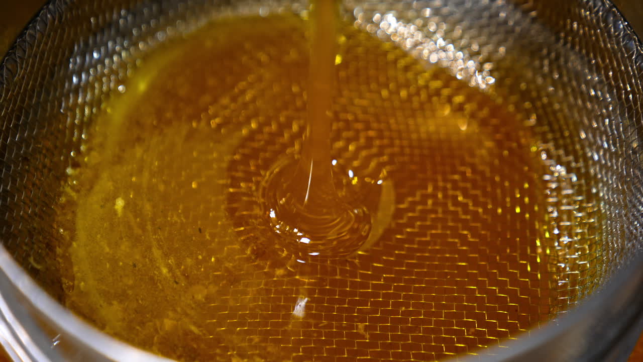 Golden flow of fresh honey leaking from machine. Honey pouring on the metal sieve for filtering from pieces of wax. Close up.