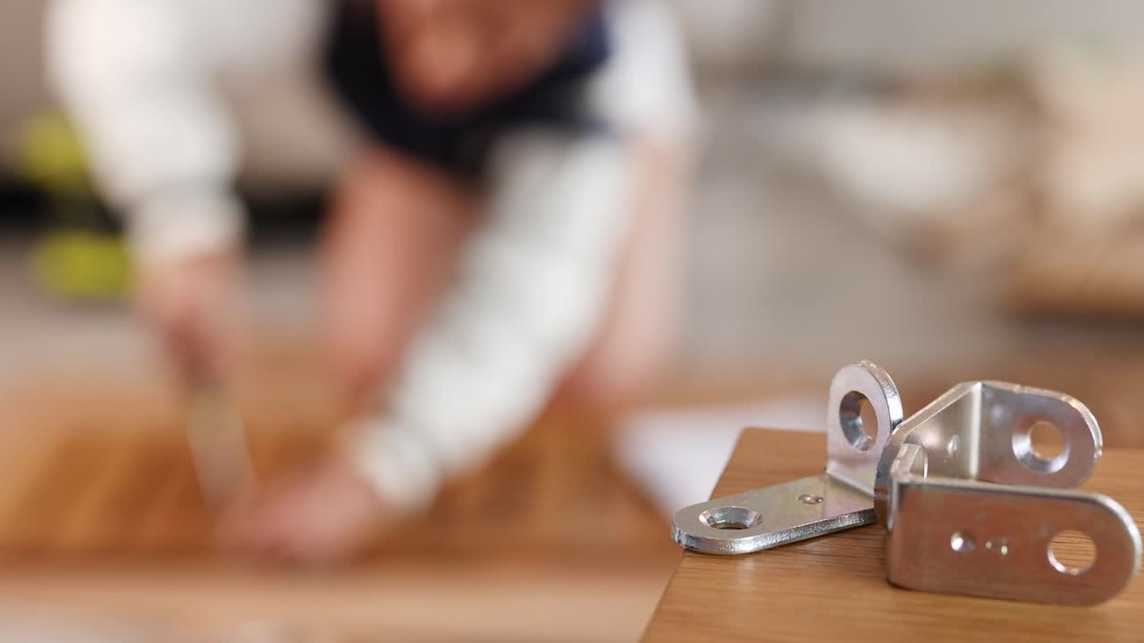 A person assembles furniture using metal brackets on a wooden surface. The background is blurred, focusing on the hardware