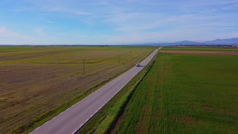 furgoneta blanca conduciendo por una sola carretera recta rodeada de verdes prados y campos de cultivo con montañas al fondo