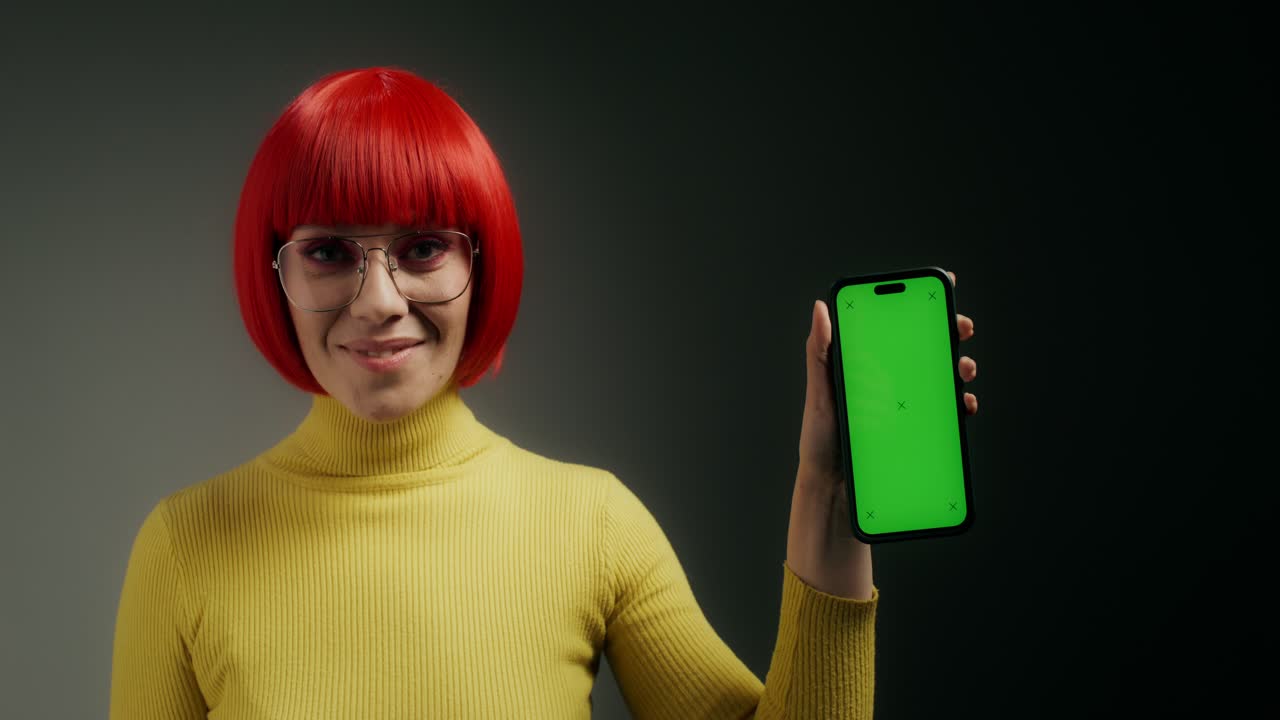 mujer con peluca roja y suéter amarillo sosteniendo un teléfono de pantalla verde