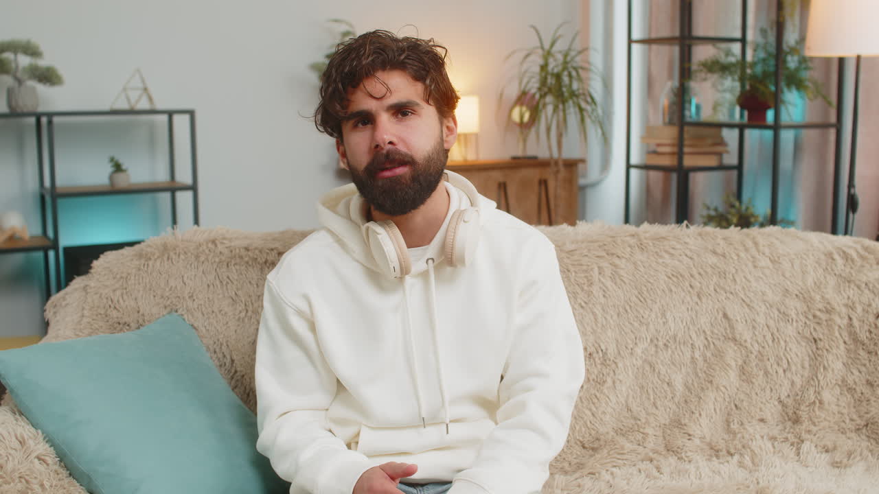 joven vlogger feliz sonriendo amigablemente a la cámara y agitando la mano mientras está sentado en el sofá en casa