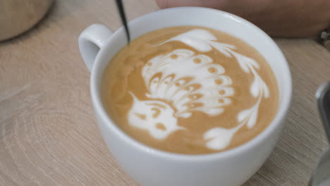 on the table is a cup of coffee with foam and the man draws on the foam pattern