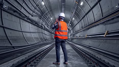 full body back view of asian male engineer with safety helmet talking on smartphone while standing in underground subway tunnel