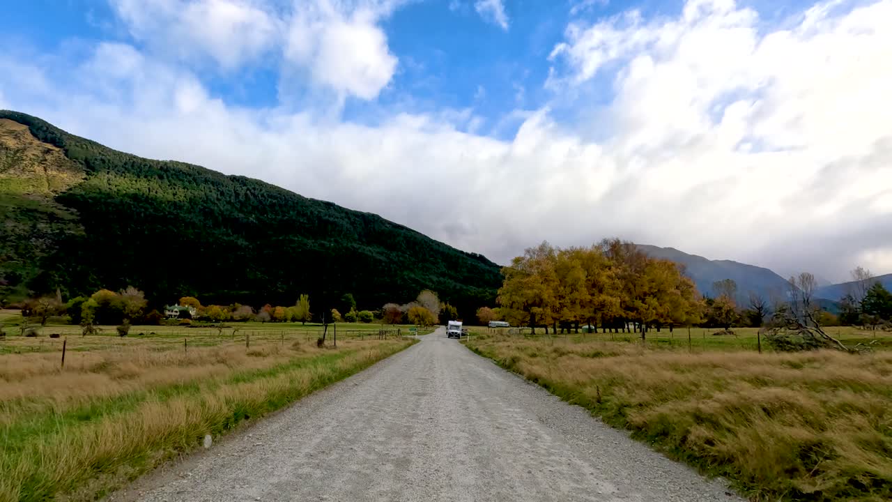 Vehicle drives toward camera on scenic gravel road, surrounded by autumn trees and mountain landscape