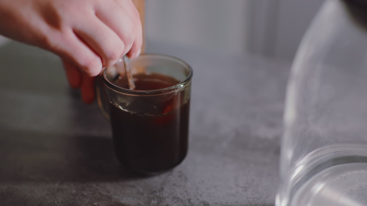 Primo piano di una mano che solleva un cucchiaino da una tazza di caffè dopo aver mescolato, superficie del liquido scuro caldo che si calma, leggere increspature che svaniscono, semplice momento della preparazione di una bevanda in cucina
